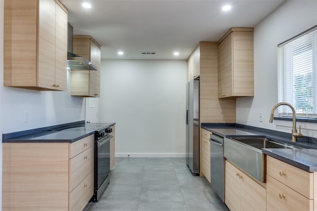 kitchen with sink, light brown cabinets, wall chimney range hood, dark stone counters, and appliances with stainless steel finishes