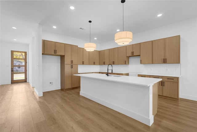 kitchen with light wood-type flooring, tasteful backsplash, a kitchen island with sink, sink, and hanging light fixtures