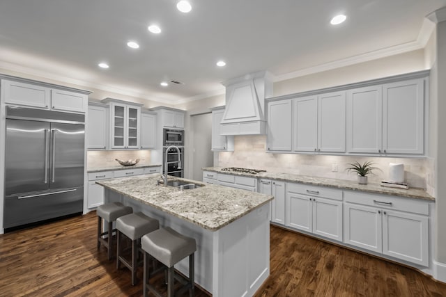 kitchen featuring built in appliances, decorative backsplash, a center island with sink, and premium range hood