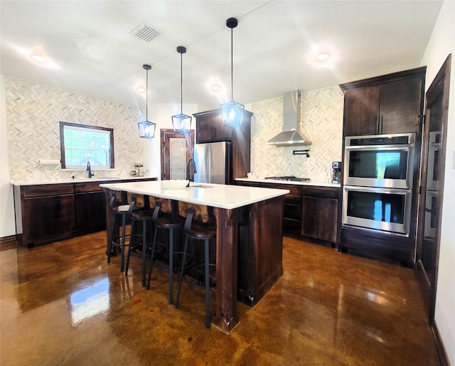 kitchen featuring a kitchen island with sink, wall chimney exhaust hood, appliances with stainless steel finishes, tasteful backsplash, and decorative light fixtures