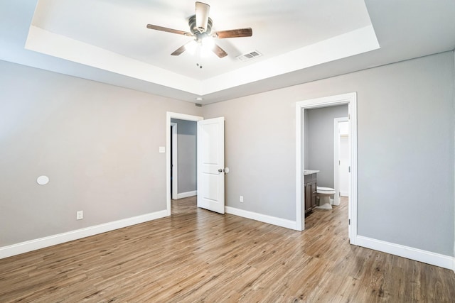 unfurnished bedroom featuring a tray ceiling, connected bathroom, ceiling fan, and light hardwood / wood-style flooring