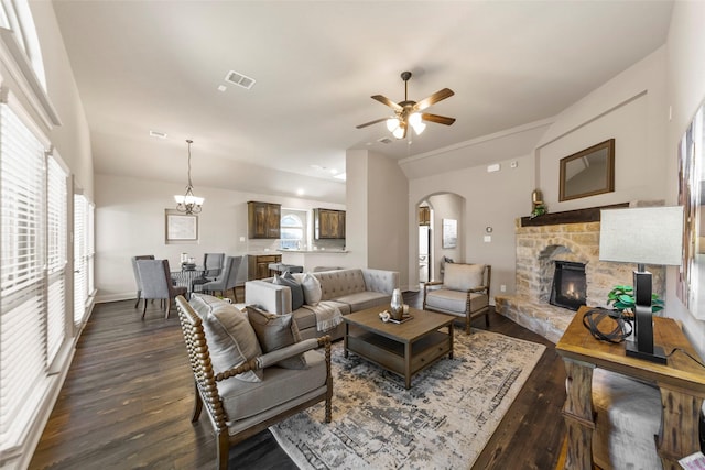 living room with ceiling fan with notable chandelier, dark wood-type flooring, and a stone fireplace