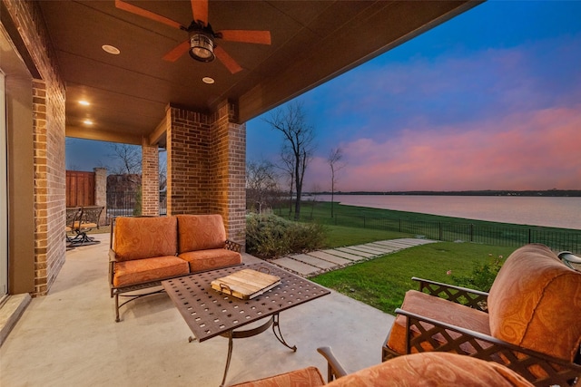 patio terrace at dusk featuring ceiling fan, a water view, an outdoor living space, and a yard