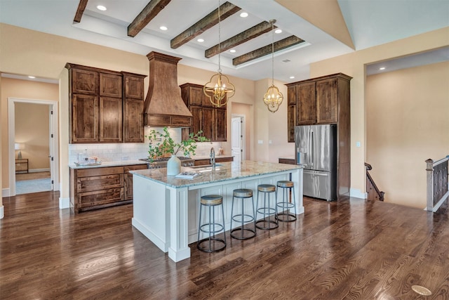 kitchen featuring light stone countertops, stainless steel refrigerator with ice dispenser, premium range hood, a kitchen island with sink, and pendant lighting