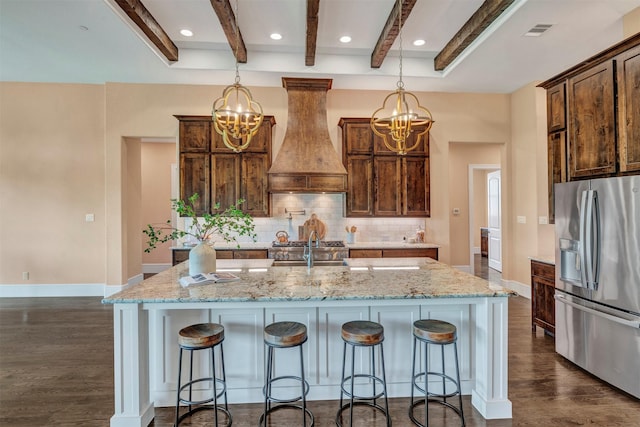 kitchen featuring pendant lighting, stainless steel fridge, custom range hood, and a kitchen island with sink