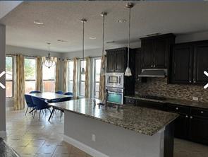 kitchen featuring tasteful backsplash, light stone counters, decorative light fixtures, oven, and an island with sink