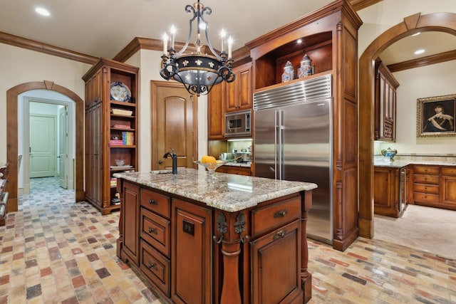 kitchen with light stone counters, a notable chandelier, built in appliances, pendant lighting, and a kitchen island with sink