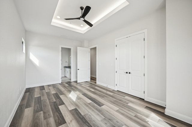 unfurnished bedroom with light wood-type flooring, a tray ceiling, ceiling fan, connected bathroom, and a closet