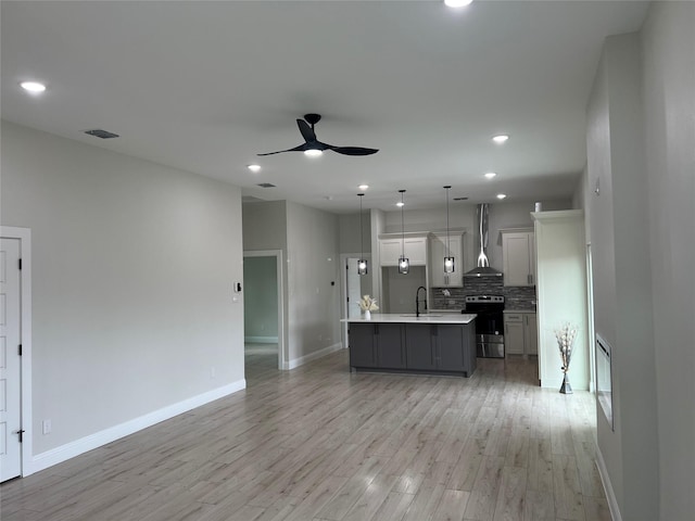 kitchen featuring light wood-type flooring, wall chimney range hood, pendant lighting, stainless steel range with electric cooktop, and an island with sink