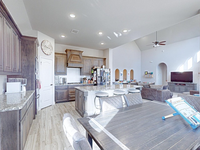 kitchen with a kitchen island with sink, appliances with stainless steel finishes, light stone counters, decorative backsplash, and wall chimney exhaust hood