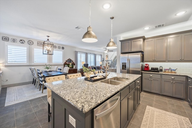 kitchen featuring light stone counters, ceiling fan with notable chandelier, stainless steel appliances, a kitchen island with sink, and hanging light fixtures