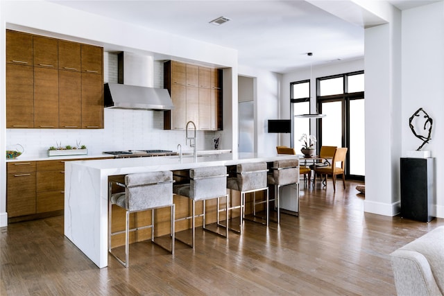 kitchen with decorative backsplash, a kitchen island with sink, a breakfast bar area, and wall chimney range hood