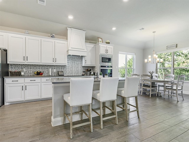 kitchen featuring stainless steel appliances, decorative light fixtures, a center island with sink, white cabinets, and custom range hood