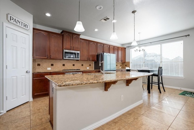 kitchen featuring appliances with stainless steel finishes, hanging light fixtures, backsplash, light stone countertops, and an island with sink