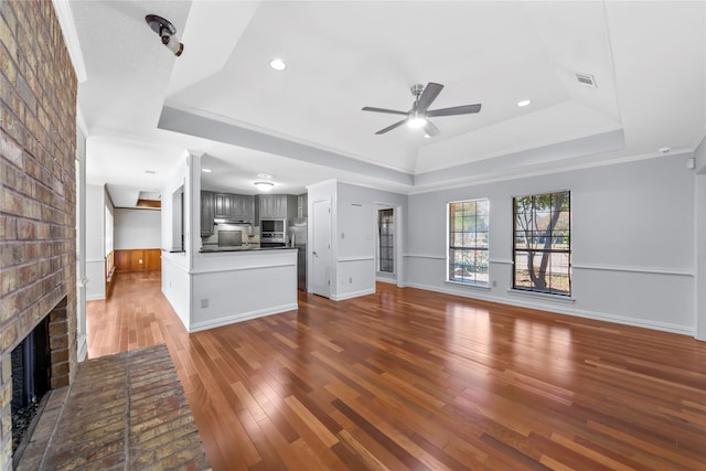 unfurnished living room featuring a brick fireplace, a raised ceiling, ceiling fan, and wood-type flooring