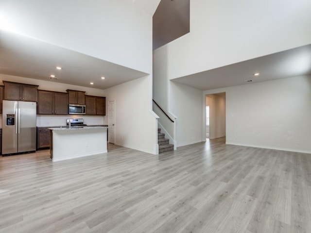 kitchen with tasteful backsplash, a towering ceiling, an island with sink, appliances with stainless steel finishes, and light wood-type flooring