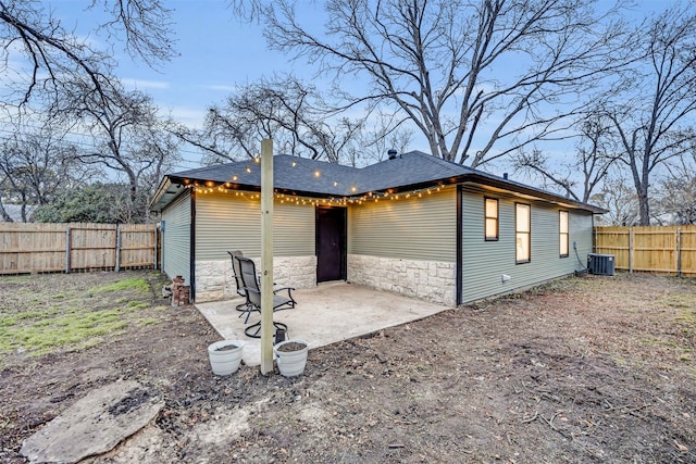 rear view of property featuring central AC unit, a fenced backyard, a shingled roof, stone siding, and a patio area