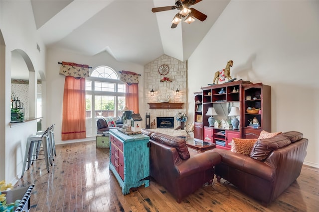 living room featuring wood-type flooring, high vaulted ceiling, a stone fireplace, and ceiling fan