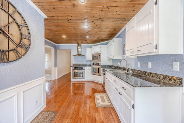 kitchen with wood ceiling, stainless steel appliances, wall chimney range hood, sink, and white cabinets