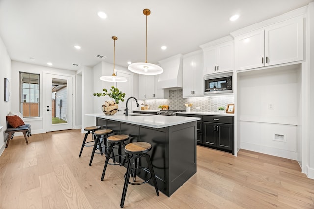 kitchen featuring pendant lighting, custom exhaust hood, white cabinetry, an island with sink, and built in microwave