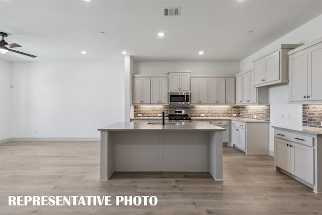 kitchen with appliances with stainless steel finishes, ceiling fan, a kitchen island with sink, sink, and light hardwood / wood-style flooring