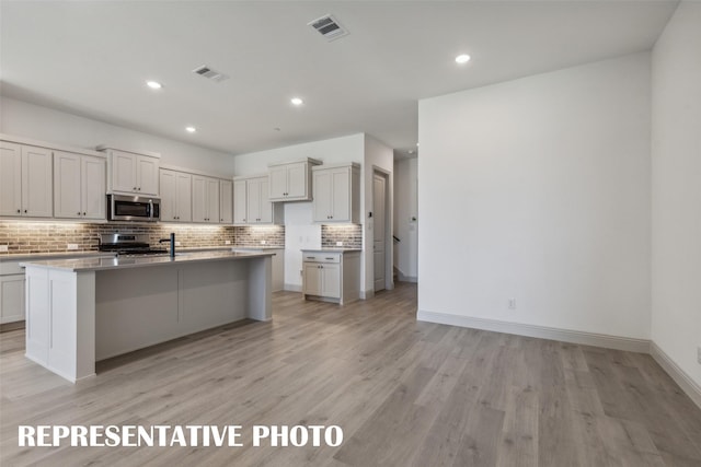 kitchen with backsplash, a kitchen island with sink, white cabinets, light hardwood / wood-style flooring, and appliances with stainless steel finishes