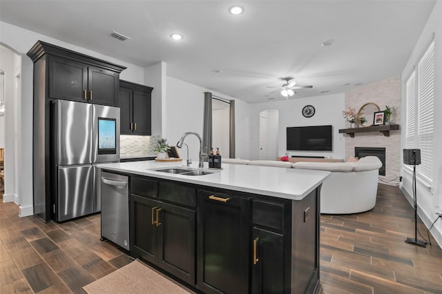 kitchen featuring stainless steel appliances, ceiling fan, a kitchen island with sink, sink, and a fireplace