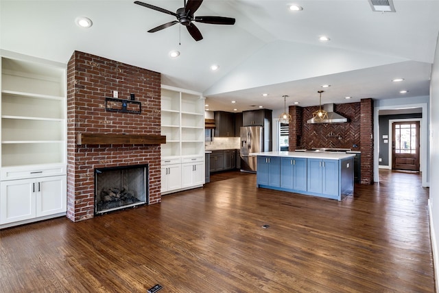 kitchen featuring stainless steel fridge, a kitchen island with sink, wall chimney range hood, a fireplace, and hanging light fixtures