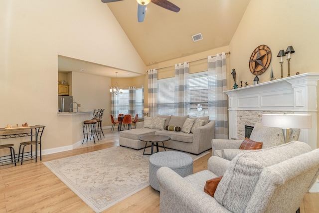 living room featuring a stone fireplace, ceiling fan with notable chandelier, high vaulted ceiling, and light hardwood / wood-style flooring