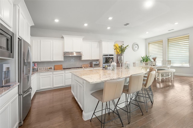 kitchen with dark wood-style flooring, stainless steel appliances, tasteful backsplash, under cabinet range hood, and a kitchen breakfast bar