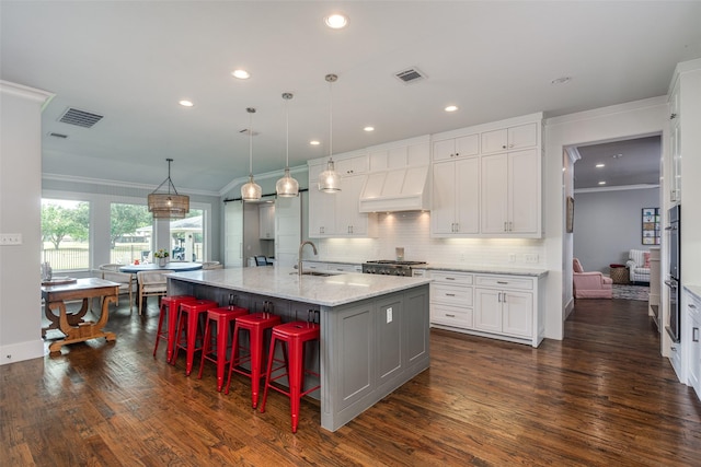 kitchen featuring light stone counters, premium range hood, a large island with sink, pendant lighting, and white cabinets