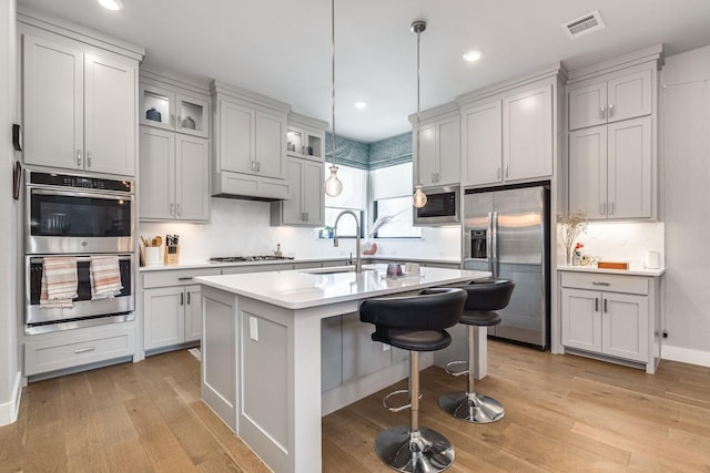 kitchen with sink, hanging light fixtures, stainless steel appliances, light hardwood / wood-style flooring, and a center island with sink