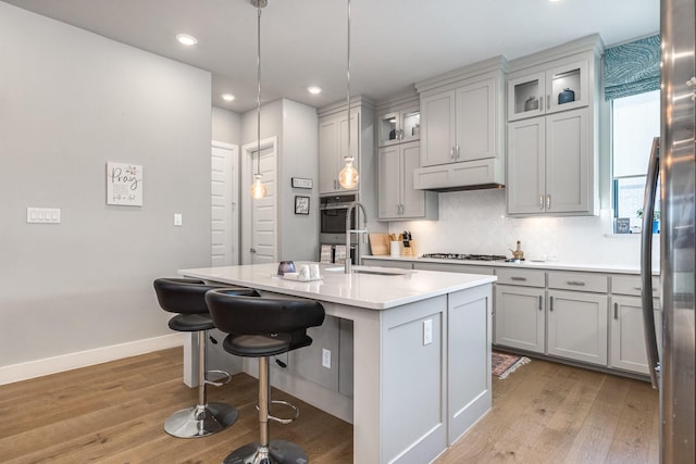 kitchen with a kitchen island with sink, decorative backsplash, light wood-type flooring, decorative light fixtures, and stainless steel appliances