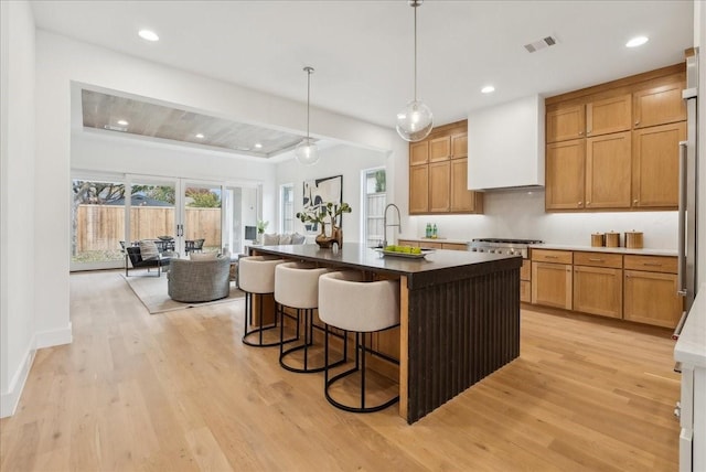 kitchen with pendant lighting, high end range, premium range hood, a kitchen island with sink, and light hardwood / wood-style floors