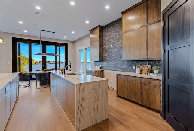 kitchen featuring backsplash, light hardwood / wood-style floors, decorative light fixtures, a kitchen island with sink, and a water view