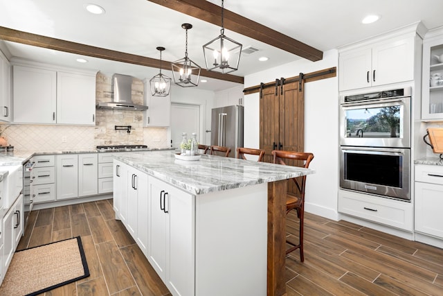 kitchen featuring stainless steel appliances, wall chimney range hood, beam ceiling, a barn door, and white cabinetry