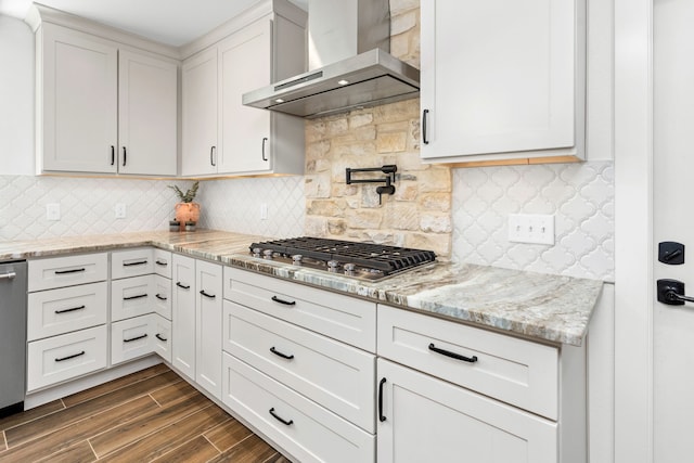 kitchen featuring wall chimney exhaust hood, light stone countertops, appliances with stainless steel finishes, tasteful backsplash, and white cabinetry