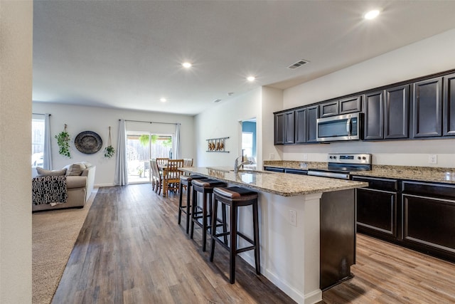 kitchen with appliances with stainless steel finishes, light stone counters, a breakfast bar, light hardwood / wood-style flooring, and an island with sink