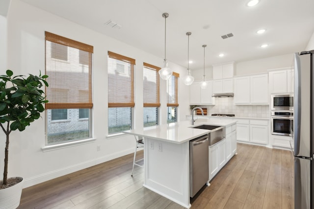 kitchen featuring stainless steel appliances, an island with sink, hanging light fixtures, white cabinets, and backsplash