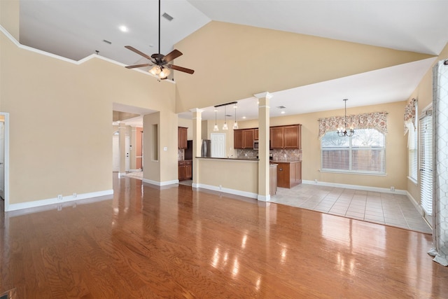 unfurnished living room with ornate columns, high vaulted ceiling, ceiling fan with notable chandelier, and light hardwood / wood-style floors