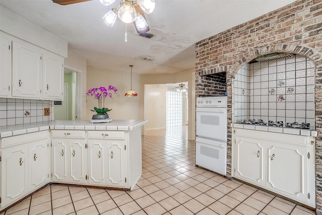 kitchen featuring white double oven, stainless steel gas cooktop, tile countertops, a textured ceiling, and white cabinets