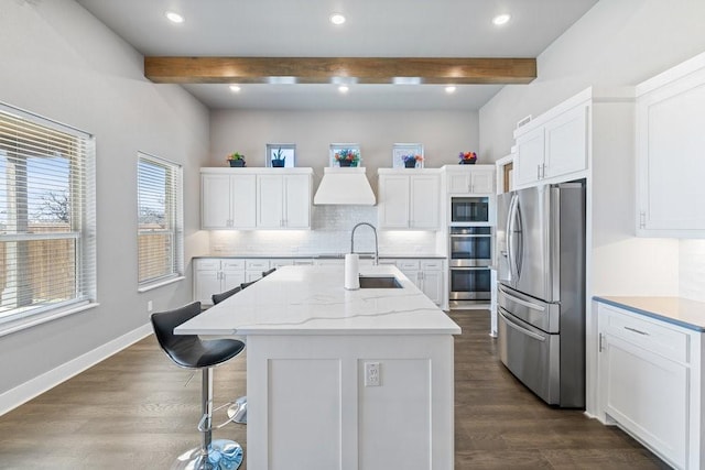 kitchen featuring appliances with stainless steel finishes, a kitchen island with sink, white cabinets, beam ceiling, and backsplash