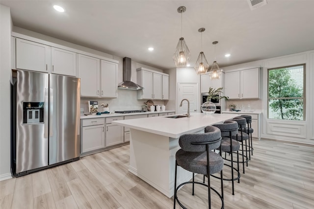 kitchen featuring pendant lighting, a center island with sink, wall chimney range hood, sink, and stainless steel refrigerator with ice dispenser