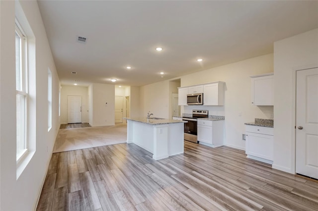 kitchen with light stone countertops, light wood-type flooring, stainless steel appliances, a center island with sink, and white cabinetry