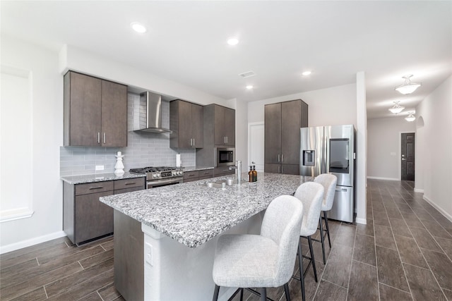 kitchen with appliances with stainless steel finishes, backsplash, light stone counters, wall chimney range hood, and an island with sink