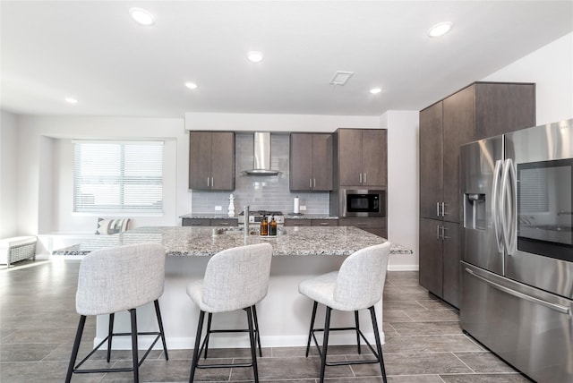 kitchen with built in microwave, wall chimney range hood, stainless steel fridge, an island with sink, and a breakfast bar area