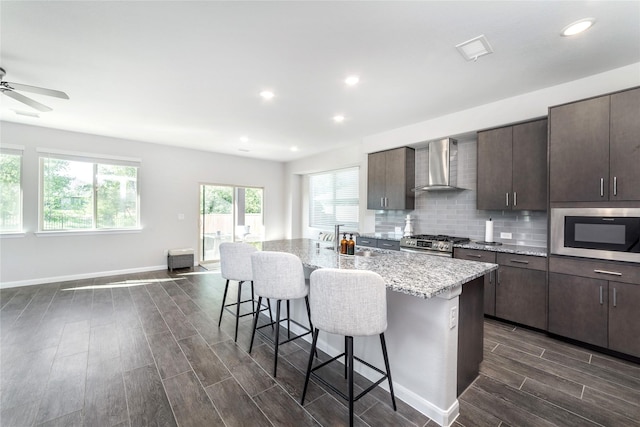 kitchen featuring stainless steel gas range oven, built in microwave, wall chimney range hood, a center island with sink, and a breakfast bar area