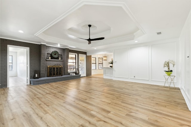 unfurnished living room featuring ceiling fan, a brick fireplace, a raised ceiling, light hardwood / wood-style flooring, and ornamental molding