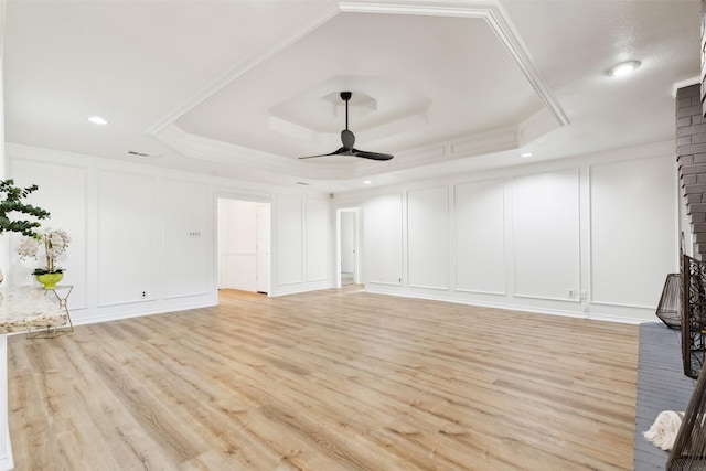 unfurnished living room with a brick fireplace, ceiling fan, a tray ceiling, light wood-style floors, and a decorative wall