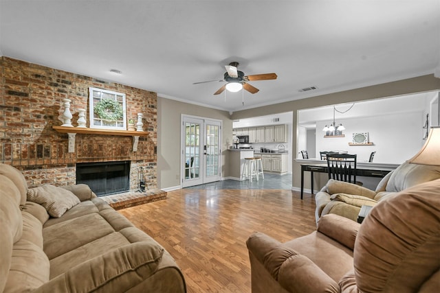 living room featuring french doors, ceiling fan with notable chandelier, crown molding, a brick fireplace, and wood-type flooring